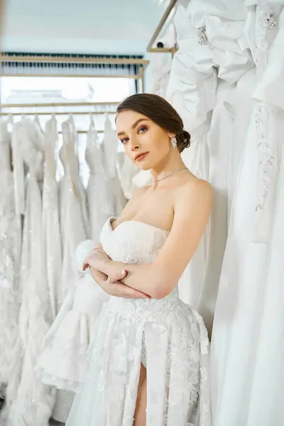A young brunette bride stands among a rack of dresses in a wedding salon, choosing her perfect gown.