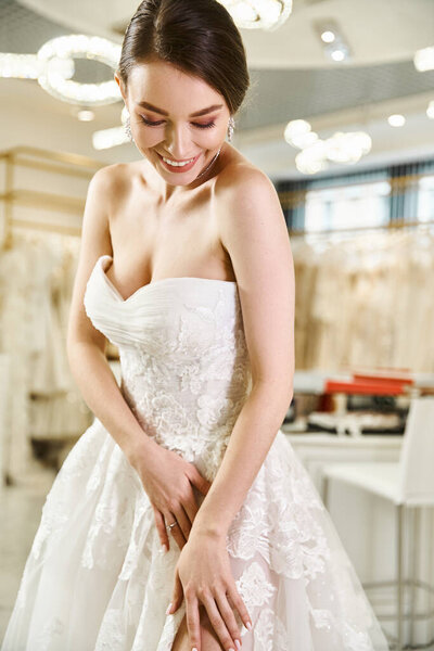 A young brunette bride is smiling happily in a white dress inside a wedding salon.