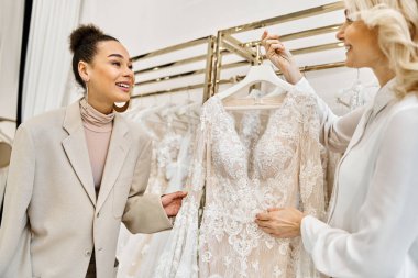 young women admiring a wedding dress on a rack in a bridal shop, assisted by a shop attendant. clipart