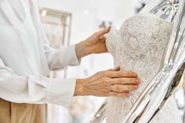 Orta yaşlı bir alışveriş asistanı, bir düğün salonundaki şık elbiselerin önünde kendinden emin bir şekilde durur..