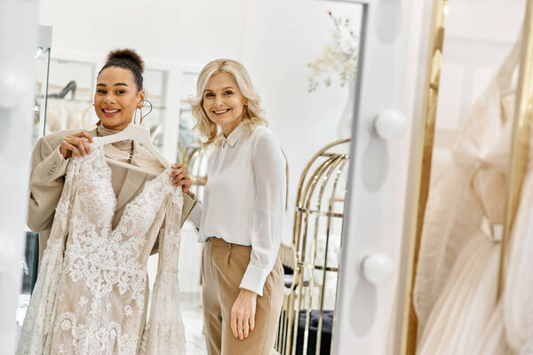 Two women, a young beautiful bride and a shop assistant, stand side by side, admiring themselves in a mirror.