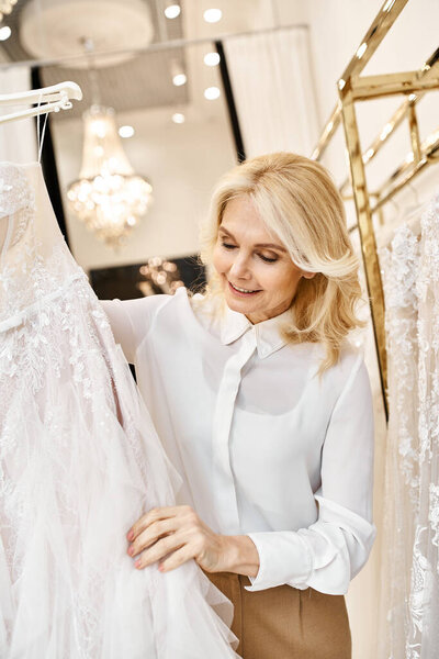 A middle-aged beautiful shopping assistant browses wedding dresses on rack in a bridal salon.
