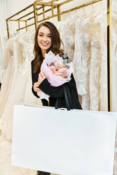 A young brunette bride holds a bouquet in front of a rack of wedding dresses in a bridal salon.