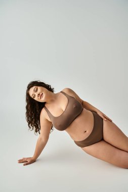 Kıvırcık kahverengi saçlı, gri arka planda poz veren ve koluna yaslanan baştan çıkarıcı kıvrımlı kadın.