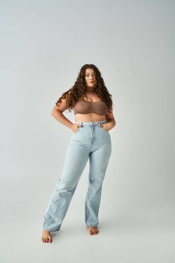 plus size woman in brown bra and blue jeans with curly hair posing against grey background clipart