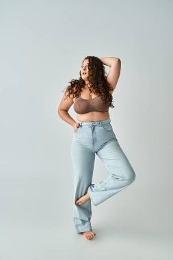 attractive curvy woman in brown bra and blue jeans posing with bent leg and putting hand behind head clipart
