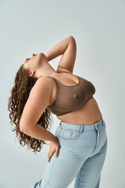 plus size young woman in brown bra and blue jeans with curly hair putting head to behind with hand clipart