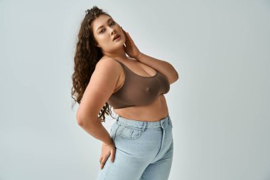 attractive plus size girl in brown bra and blue jeans posing and touching with hand to face clipart