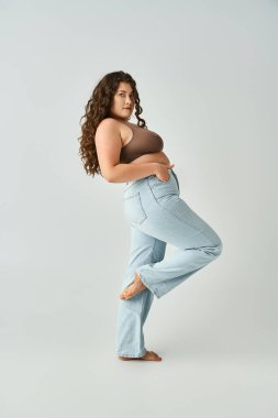 pretty curvy woman in brown bra and blue jeans leaning to behind with bent leg on grey background clipart