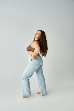 seductive plus size young woman in brown bra and blue jeans putting foot forward on grey background clipart