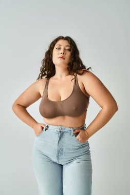 charismatic curvy girl in brown bra and blue jeans posing with hands in pockets on grey background clipart