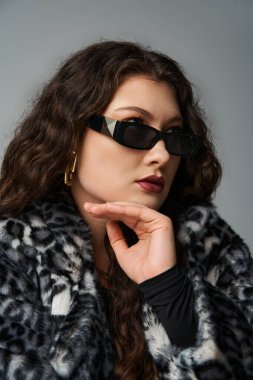 portrait of glamourous curvy woman in sunglasses looking to side with chin on hand on grey clipart