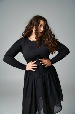 Çekici ve cüsseli genç kız elleri belinde gri arka planda taraf arıyor.