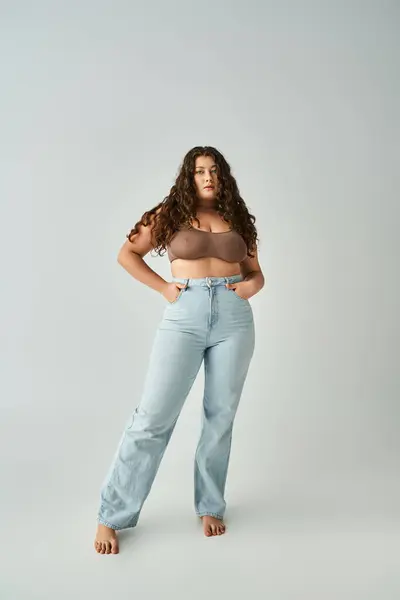 stock image plus size woman in brown bra and blue jeans with curly hair posing against grey background