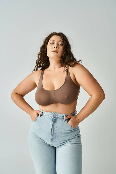charismatic curvy girl in brown bra and blue jeans posing with hands in pockets on grey background