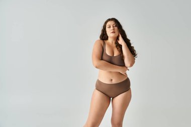 alluring plus size young woman in brown lingerie with curly hair posing with arms clipart