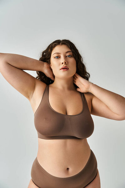 соблазнительная пышная девушка в коричневом белье с вьющимися волосами, касающимися рук к шее и смотрящая в камеру