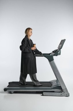 appealing graceful woman in stylish black coat with trendy glasses posing on treadmill with coffee clipart