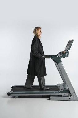 attractive sophisticated woman in fashionable black coat posing on treadmill on gray background clipart