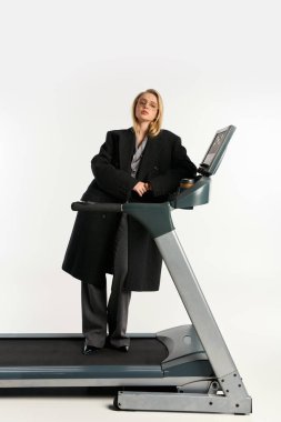 attractive graceful woman in stylish glasses and coat posing on treadmill and looking at camera clipart