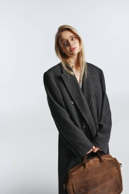 appealing graceful woman with blonde hair in black stylish coat holding brown bag and looking away clipart