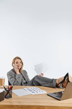 joyous businesswoman in smart gray suit with stylish glasses talking by phone while sitting at desk clipart