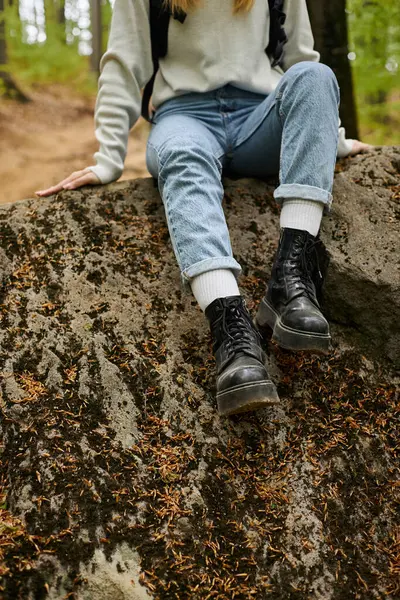 森の岩の上に座っているジーンズとハイキングブーツを身に着けている女性のハイカーの足のクロップされたイメージ — ストック写真