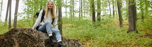 stock image Smiling pretty blonde woman hiker sitting on a rock in forest and looking away, banner