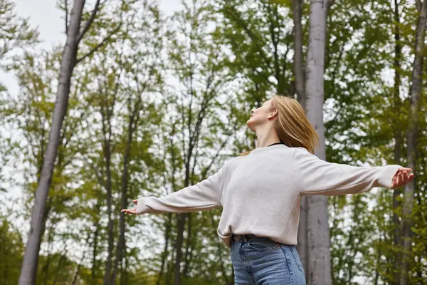 Energetic female blond forest explorer in jeans and sweater standing in serene setting, freedom