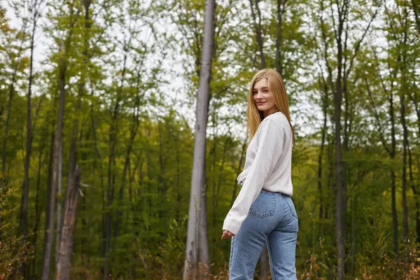 笑顔の金髪の女性は平和な森を歩いている間に周りを見回し カメラを見ています — ストック写真