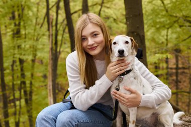 Gülümseyen sarışın kadın, orman gezisini durdururken köpeğine sarılıyor ve kameraya bakıyor.