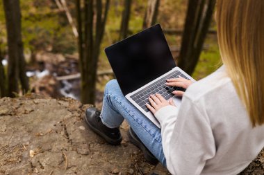 Back view of a blonde woman sitting on a large rock in the forest, using a laptop for remote work clipart