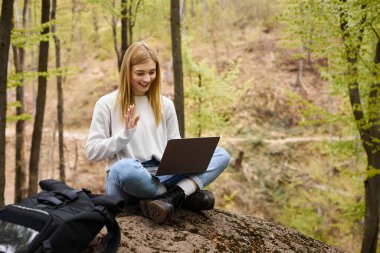 Ormanda sırt çantalı genç sarışın bir kadın dizüstü bilgisayarlı bir kayanın üzerinde oturuyor ve video görüşmeleri yapıyor.