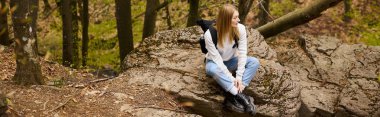 Young woman hiker with backpack sitting at halt on rocky cliff in forest looking away, banner clipart