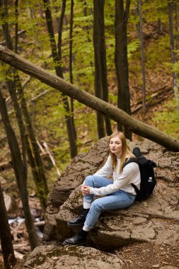 Relaxed woman traveler with backpack sitting at halt on rocky cliff in forest scenery clipart