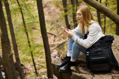 Side view of woman holding phone while resting in the forest. Hiking and sightseeing concept clipart