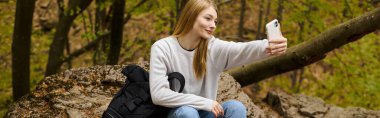 blonde young woman taking selfie sitting on rock at halt in the forest while hiking, banner clipart