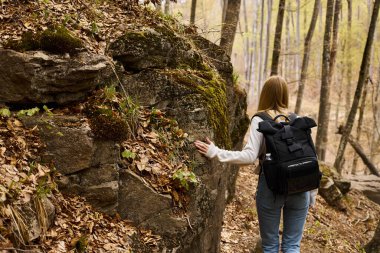 Back view of blonde female traveler wearing backpack on hiking trip and walking near rocky cliff clipart