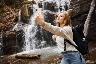 Sarışın genç kadın dağ şelalesinin yakınında yürüyüş yaparken selfie çekiyor.