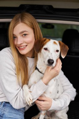 Gülümseyen mutlu kadın köpeğini kucaklıyor. Ormanda arabanın arkasında oturuyor ve kameraya bakıyor.