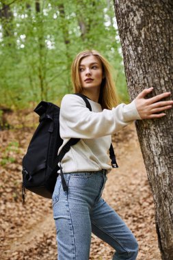 Relaxed blonde woman hiking and walking in green forest touching trees turning for direction clipart