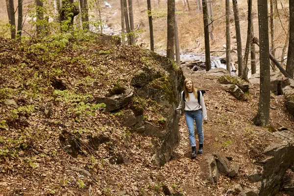 アクティブな若い金髪の女性の旅行者は ハイキング旅行でバックパックを着て 森の川の近くを歩いています — ストック写真