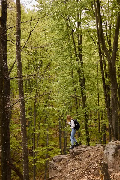 徒步旅行时穿着背包 手持电话在岩石悬崖上行走的好奇金发女人的侧影 — 图库照片