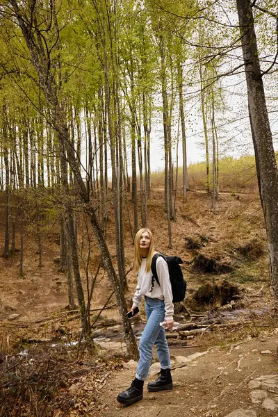 水のボトルを持っている森を渡るセーターとジーンズを身に着けている若い女性旅行者 — ストック写真