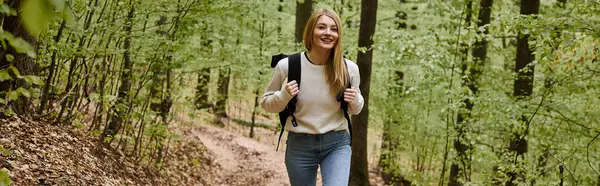 Happy hiking blonde woman wearing sweater and backpack walking in forest scenery in woods, banner