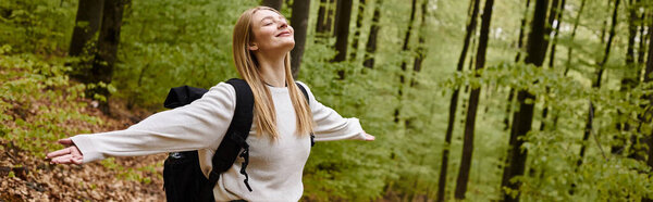 Smiling relaxed blonde woman hiker wearing sweater and backpack with arms  open in forest, banner