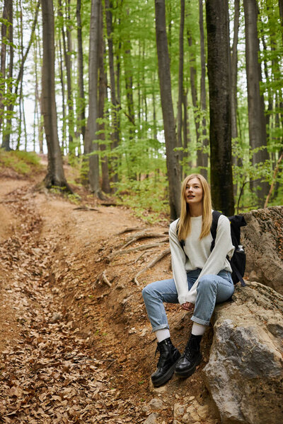 Улыбающаяся блондинка-туристка в рюкзаке и отдыхающая на камнях, сидя в глубоком лесу