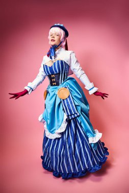 Mavi şapkalı ve canlı giyinmiş, pembe arka planda kameraya bakan çekici bir kadın Cosplayer.