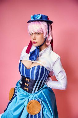 Çekici bayan Cosplayer mavi şapkalı ve canlı giyinmiş pembe arka planda kameraya bakıyor.