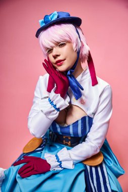 Baştan çıkarıcı seksi bayan Cosplayer mavi şapkalı ve canlı giyinmiş pembe arka planda kameraya bakıyor.